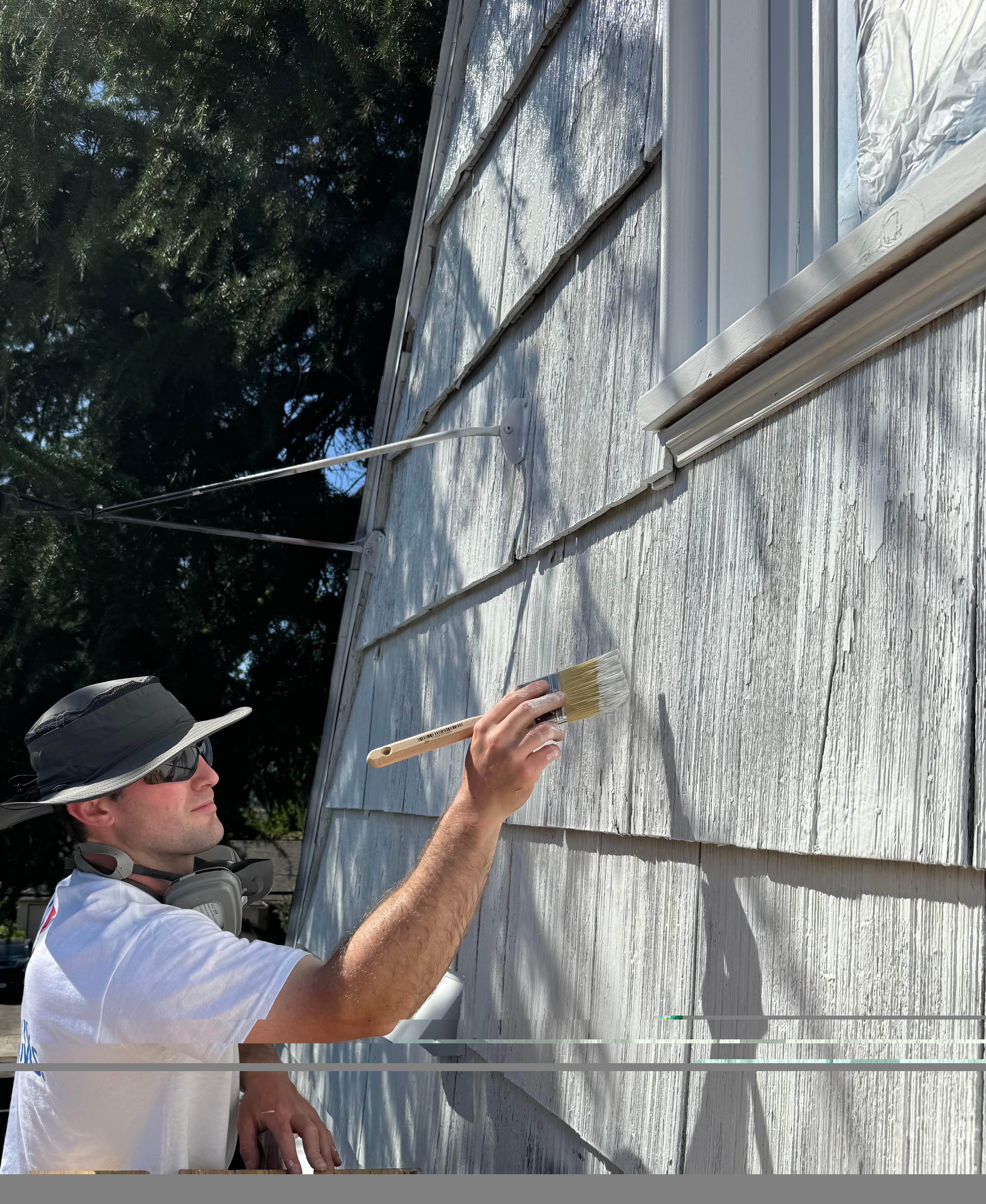 Man wearing a hat and sunglasses applies white paint to grey wooden house shingles.